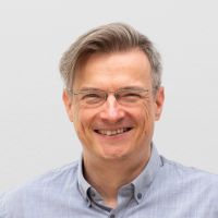 Tobias Bonnke -Solution Architect : Homme souriant avec des cheveux gris et lisses peignés sur le côté et de fines lunettes carrées grises dont seule la moitié supérieure est encadrée.